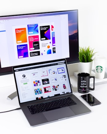 A modern workspace featuring a MacBook Pro with open design software and a large monitor displaying colorful design reports. A black mug, a potted plant, and a smartphone are also visible on the white desk. A Starbucks cup is placed near the monitor.