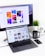 A modern workspace featuring a MacBook Pro with open design software and a large monitor displaying colorful design reports. A black mug, a potted plant, and a smartphone are also visible on the white desk. A Starbucks cup is placed near the monitor.