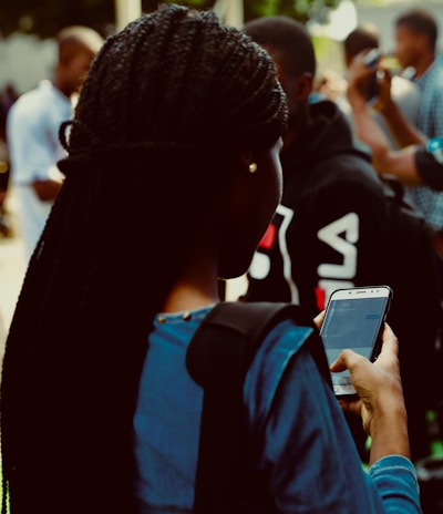 A person with braided hair is using a smartphone. They are wearing a blue top and are surrounded by a crowd in an outdoor setting. The smartphone screen is visible with a messaging app open.