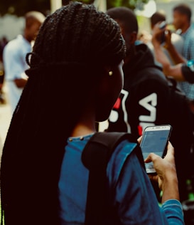 A person with braided hair is using a smartphone. They are wearing a blue top and are surrounded by a crowd in an outdoor setting. The smartphone screen is visible with a messaging app open.