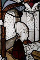 A stained glass depiction of a religious figure wearing a mitre and holding a scroll inside an architectural setting with ornate features. The artwork includes intricate window designs and detailed facial expressions.