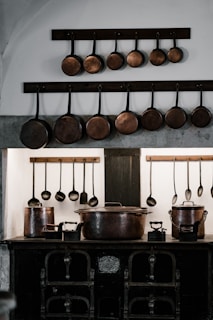 A rustic kitchen setting features a collection of copper pans and ladles hanging on a wall above a vintage stove. The arrangement is symmetrical, with the pans varying in size. Below, two large copper pots and cast iron trivets are placed on the stove, making the scene reminiscent of an old-world kitchen.