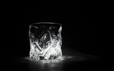 A single, intricately designed glass is placed on a dark surface, illuminated from below. The light enhances the textured surface of the glass, creating intricate patterns and reflections.