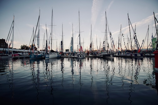 A marina filled with numerous sailboats, each with tall masts reflecting in the calm water. The sky is clear with streaks of clouds. Flags are hanging from many of the boats, indicating a festive or competitive event. The water is smooth, adding to the serene atmosphere.
