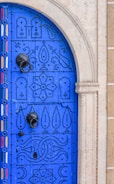 A vibrant blue door adorned with intricate black studs forming patterns, including geometric shapes and abstract designs. The door features a large black metal door knocker and is set within a light stone archway. Stripes of red, yellow, and white add an additional decorative element along the edge.