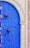 A vibrant blue door adorned with intricate black studs forming patterns, including geometric shapes and abstract designs. The door features a large black metal door knocker and is set within a light stone archway. Stripes of red, yellow, and white add an additional decorative element along the edge.