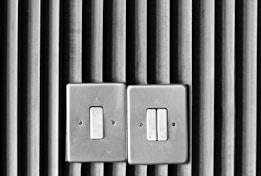 Two metal light switches are mounted on a wall with vertical wooden slats. The design is simplistic, with a textured backdrop providing contrast to the smooth, metallic surface of the switches.