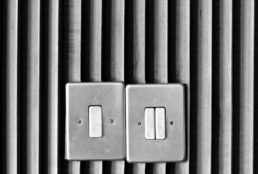 Two metal light switches are mounted on a wall with vertical wooden slats. The design is simplistic, with a textured backdrop providing contrast to the smooth, metallic surface of the switches.