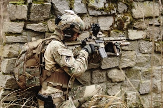 A soldier in camouflage gear is crouched against a moss-covered stone wall, holding an assault rifle. The soldier is wearing a helmet with a mounted camera and a tactical backpack, appearing to be in an outdoor, possibly wooded, environment.
