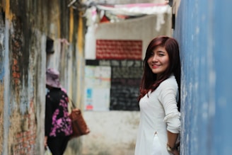 A young woman with long hair is leaning against a blue wall, smiling. She is wearing a white dress. In the background, an alley with worn walls is visible, and another person, partially obscured, is walking away while carrying a brown bag.