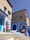 The scene features traditional Moroccan architecture with buildings painted in shades of blue. Two women wearing colorful traditional clothing and hats are present. One is standing near a blue door, while the other is seated on a blue step, with a decorative straw hat placed on the foreground step. The setting is sunlit, with a clear blue sky overhead.