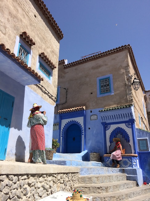 The scene features traditional Moroccan architecture with buildings painted in shades of blue. Two women wearing colorful traditional clothing and hats are present. One is standing near a blue door, while the other is seated on a blue step, with a decorative straw hat placed on the foreground step. The setting is sunlit, with a clear blue sky overhead.