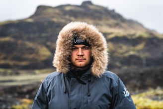 A rugged men's winter jacket with a fur-lined hood hanging on a rustic wooden fence dusted with snow.