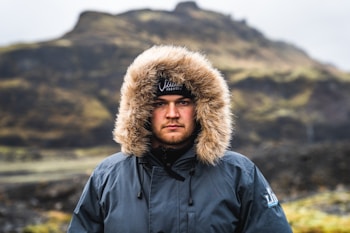 A man is wearing a winter jacket with a fur-lined hood, standing outdoors. The background features a blurred landscape with rocky hills and muted green and brown tones, suggesting a rugged, cold environment.