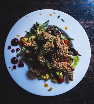 A white plate filled with a colorful salad topped with crispy fried chicken pieces. The salad includes cherry tomatoes, lettuce, and a variety of greens, with bright sauces artfully drizzled around the edge of the plate. Some dark spots of sauce provide contrast against the white background.