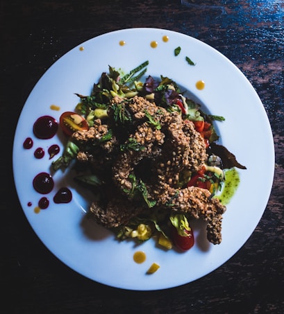 A white plate filled with a colorful salad topped with crispy fried chicken pieces. The salad includes cherry tomatoes, lettuce, and a variety of greens, with bright sauces artfully drizzled around the edge of the plate. Some dark spots of sauce provide contrast against the white background.