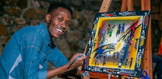 A person is smiling while painting on a canvas placed on an easel. The artwork is colorful and features abstract figures and patterns. The individual is wearing a denim shirt and appears to be enjoying the creative process.