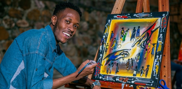 A person is smiling while painting on a canvas placed on an easel. The artwork is colorful and features abstract figures and patterns. The individual is wearing a denim shirt and appears to be enjoying the creative process.