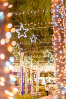 A vibrant display of colorful star-shaped lights adorns the street, creating a festive atmosphere. Strings of white and colorful lights are draped over trees, with palm fronds visible in the background. A woman and child walk along a path, enjoying the illuminated scene.