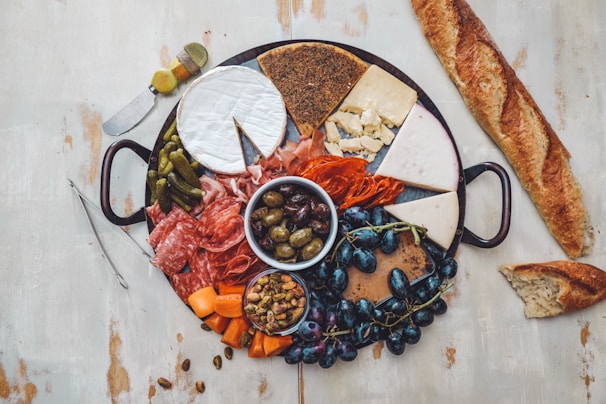 A wooden table is set with a large charcuterie board featuring an assortment of meats, cheeses, and accompaniments. Slices of cured meats like salami and prosciutto are arranged alongside wedges of various cheeses, including a wheel of Brie with a piece cut out. The board also contains small pickles, olives, grapes, and a bowl of nuts. A whole baguette and a slice are placed beside the board, and there is a small serving knife on the table.