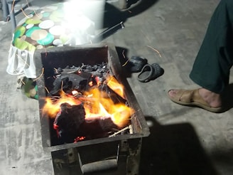 A charcoal grill with visible flames and glowing coals is placed on the ground. Nearby, a person’s foot wearing a sandal is partially visible. There are also some slippers and a cushion with a colorful circular pattern in the background.