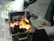 A charcoal grill with visible flames and glowing coals is placed on the ground. Nearby, a person’s foot wearing a sandal is partially visible. There are also some slippers and a cushion with a colorful circular pattern in the background.