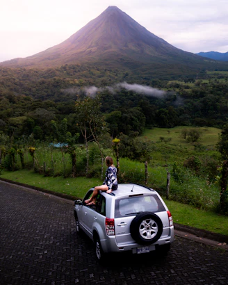 landscape photo of woman sitting on silver car looking at mountains during daytime