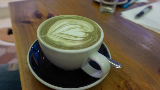 A cup of coffee with intricate latte art sits on a dark saucer. The wooden table supporting the cup has a warm, natural finish. In the background, there are a pen, paper, and a glass, slightly out of focus.