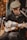 A person is sitting and playing an acoustic guitar. They are wearing a flat cap and a dark jacket, focused intently on the instrument. The setting appears to be a market or outdoor area with shelves of pottery in the background.