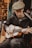 A person is sitting and playing an acoustic guitar. They are wearing a flat cap and a dark jacket, focused intently on the instrument. The setting appears to be a market or outdoor area with shelves of pottery in the background.