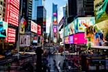 A bustling urban square filled with bright, colorful billboards and advertisements on tall buildings. The street is busy with people walking and cars driving past, with vibrant digital screens displaying various advertisements. Flags are visible, and a wide pedestrian area is central to the scene.