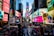 A bustling urban square filled with bright, colorful billboards and advertisements on tall buildings. The street is busy with people walking and cars driving past, with vibrant digital screens displaying various advertisements. Flags are visible, and a wide pedestrian area is central to the scene.