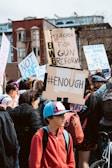 A crowd of people participating in a protest, holding signs with messages advocating for gun reform. One sign prominently reads '#ENOUGH' while others display messages like 'Teacher for Gun Reform' and 'Guns don't kill people... yes they do!'. The scene takes place in an urban setting with buildings in the background.