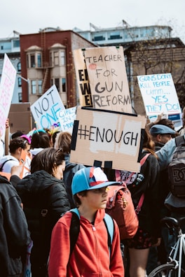 A crowd of people participating in a protest, holding signs with messages advocating for gun reform. One sign prominently reads '#ENOUGH' while others display messages like 'Teacher for Gun Reform' and 'Guns don't kill people... yes they do!'. The scene takes place in an urban setting with buildings in the background.