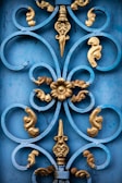 A detailed ornamental ironwork design featuring swirling patterns and a central floral motif. The decorative piece is highlighted by a combination of blue and gold colors, showcasing intricate craftsmanship.