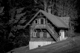 A rustic wooden cabin is nestled amidst tall trees, featuring a steeply pitched roof and a small staircase leading to an upper level. The cabin exudes a traditional and quaint charm, surrounded by a dense forest of fir trees. The black and white composition adds a timeless and serene quality to the scene.