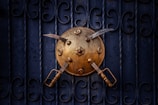 A brass shield adorned with embossed floral designs is mounted on a dark, ornately patterned metal gate. Two curved swords with wooden handles are crossed over the shield, creating a striking visual contrast between the gold and the deep blue background.