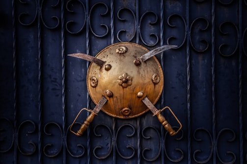 A brass shield adorned with embossed floral designs is mounted on a dark, ornately patterned metal gate. Two curved swords with wooden handles are crossed over the shield, creating a striking visual contrast between the gold and the deep blue background.
