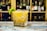 A cocktail sits elegantly in a faceted glass, garnished with a twist of lime peel, positioned on a rustic wooden bar. In the blurred background, shelves of various bottles create a warm, inviting atmosphere.
