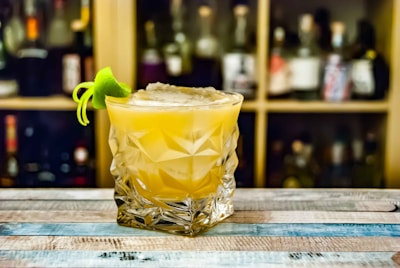 A cocktail sits elegantly in a faceted glass, garnished with a twist of lime peel, positioned on a rustic wooden bar. In the blurred background, shelves of various bottles create a warm, inviting atmosphere.