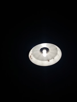 A bright light bulb is partially enclosed by a metallic or plastic lampshade. The surface of the shade shows signs of wear or dirt. The surrounding area is very dark, highlighting the light source.