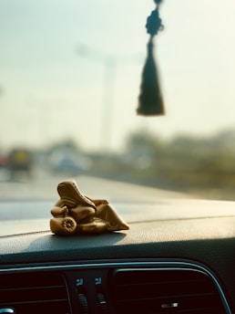 A small golden statue of Ganesha is placed on the dashboard of a car. The background shows a blurred view of a road with vehicles, and a hanging car accessory is visible from the rearview mirror.