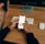 A person is using a smartphone while sitting at a wooden desk. The smartphone screen displays a social media feed with text posts and an image. In the background, there is a wireless keyboard and a wireless mouse positioned next to a monitor stand.