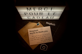 A lightbox displays the words 'MERCI POUR LE CADEAU' in illuminated letters. Below the lightbox lies a note from Unsplash expressing gratitude, placed on top of a brown envelope with a postage stamp. There is also a sticker with the Unsplash logo.