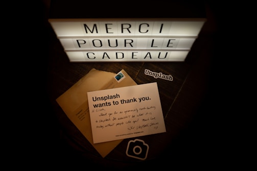 A lightbox displays the words 'MERCI POUR LE CADEAU' in illuminated letters. Below the lightbox lies a note from Unsplash expressing gratitude, placed on top of a brown envelope with a postage stamp. There is also a sticker with the Unsplash logo.