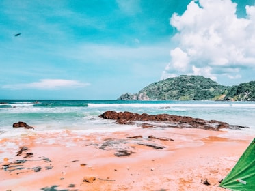 A picturesque beach with turquoise waters and reddish-brown rocks scattered across the shore. The sky is bright blue with fluffy white clouds. A green hill is visible in the distance, and the corner of a green tent is seen on the sandy foreground.