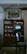 A vintage wooden cabinet with shelves filled with organized stacks of books, retro electronics, and decorative items. Above the cabinet is a framed painting depicting a group of people in colorful traditional clothing, set against a rural scene with trees and a sun. A small television displays black and white static. In the foreground, there is a dark wooden chair with patterned upholstery. The room has a rustic and nostalgic ambiance, enhanced by subdued lighting and plants in the background.