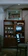 A vintage wooden cabinet with shelves filled with organized stacks of books, retro electronics, and decorative items. Above the cabinet is a framed painting depicting a group of people in colorful traditional clothing, set against a rural scene with trees and a sun. A small television displays black and white static. In the foreground, there is a dark wooden chair with patterned upholstery. The room has a rustic and nostalgic ambiance, enhanced by subdued lighting and plants in the background.