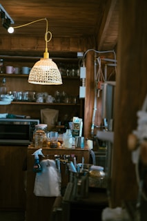 A cozy kitchen scene featuring a rustic wooden interior. Various kitchen items are placed on the counter, including a spray bottle, glass jars, a carton, and metal canisters. A mix of hanging utensils and a white cloth are visible. Overhead, a woven rattan-style light fixture is suspended, providing warm illumination.