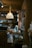 A cozy kitchen scene featuring a rustic wooden interior. Various kitchen items are placed on the counter, including a spray bottle, glass jars, a carton, and metal canisters. A mix of hanging utensils and a white cloth are visible. Overhead, a woven rattan-style light fixture is suspended, providing warm illumination.
